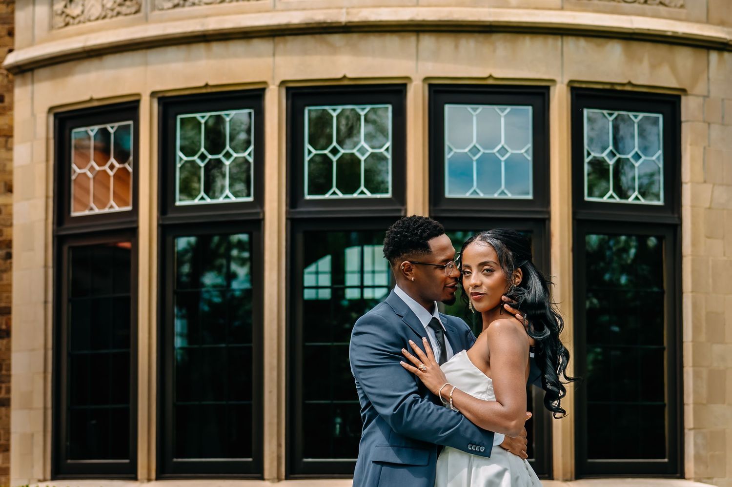 A couple in wedding attire embrace in front of ornate stained glass windows of a historic stone building.