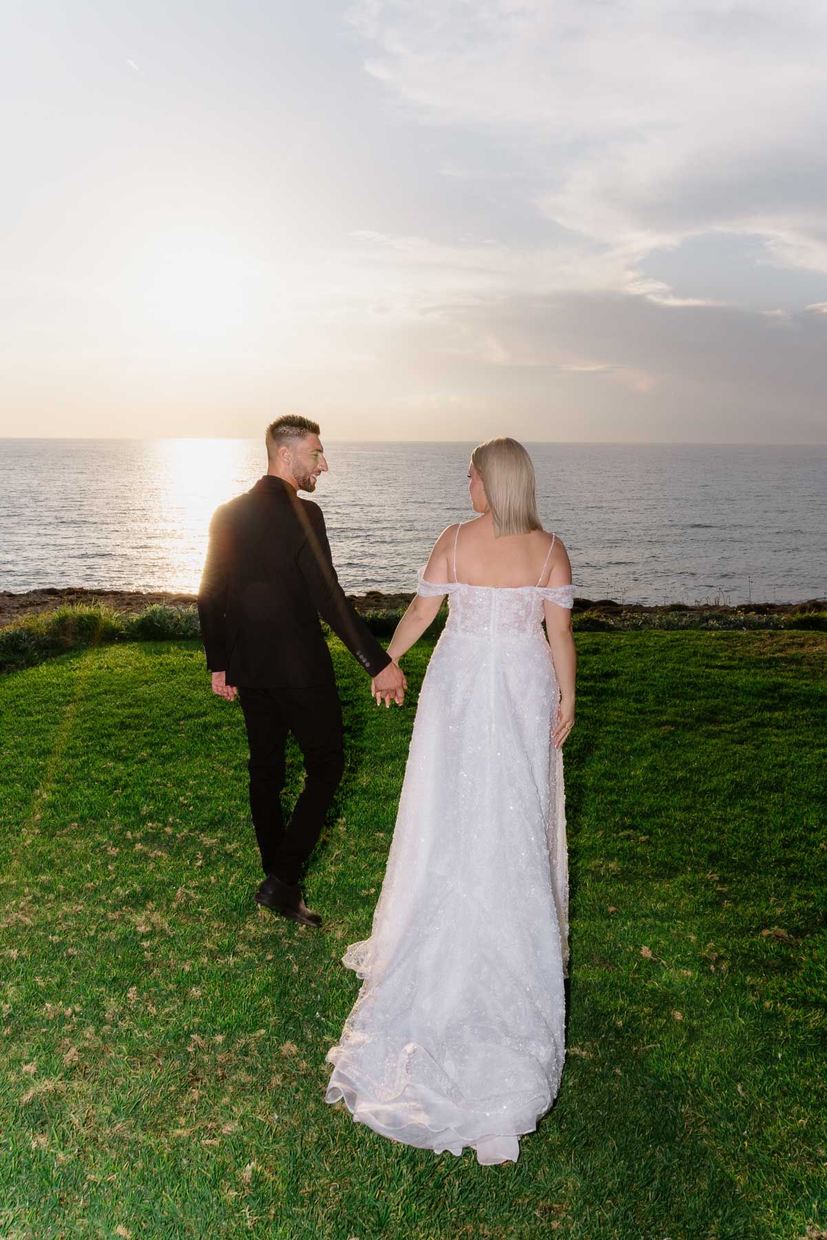 A couple in wedding attire walks along a grassy coastal cliff at sunset, creating a romantic silhouette against the ocean.
