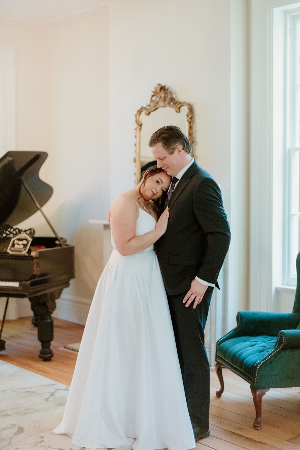 A bride and groom stands together in the Larimore Wedding Venue parlor, the bride leaning her head on the groom's shoulder.