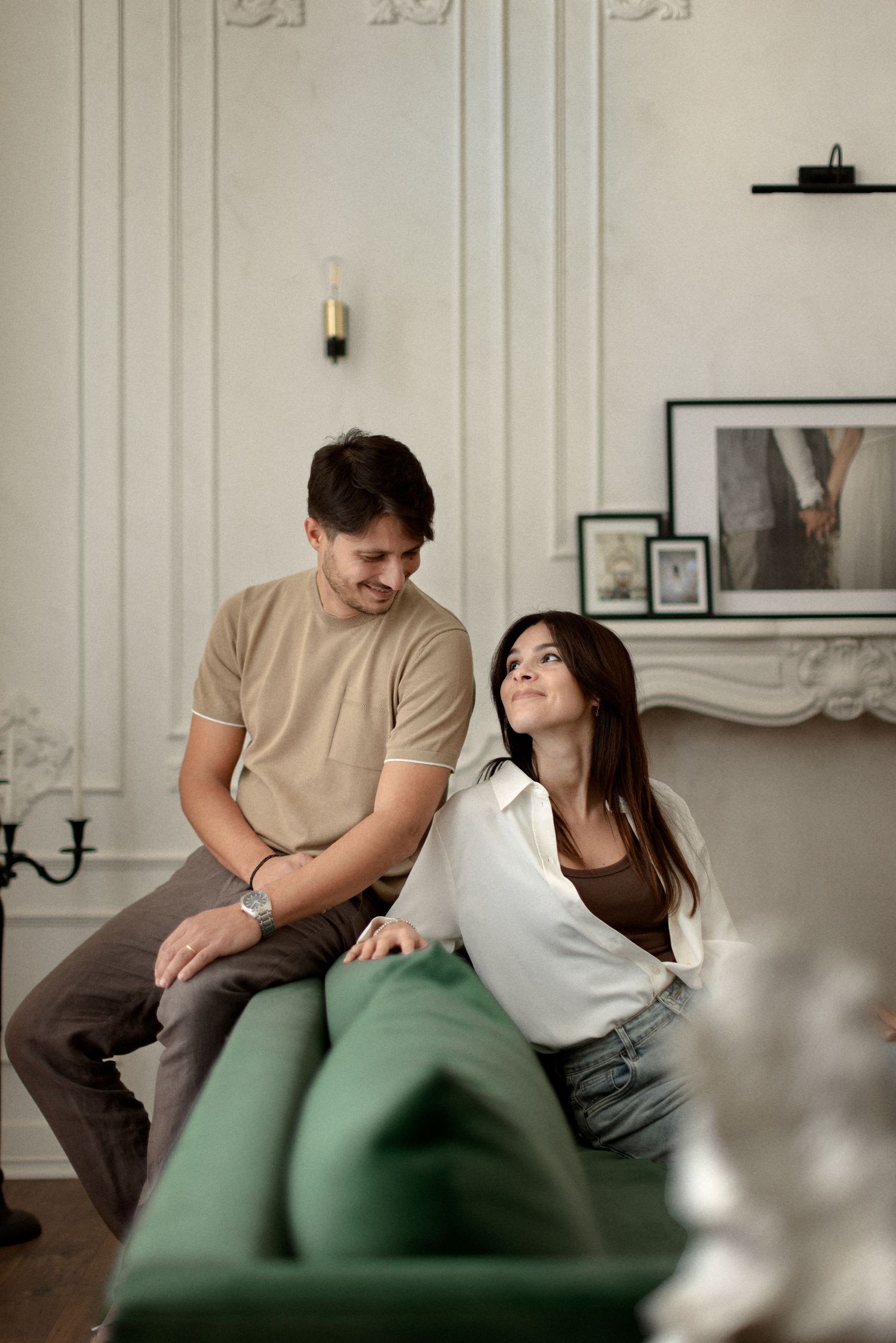 A couple relaxes together on a green couch in an elegant room with white molded walls and framed artwork.