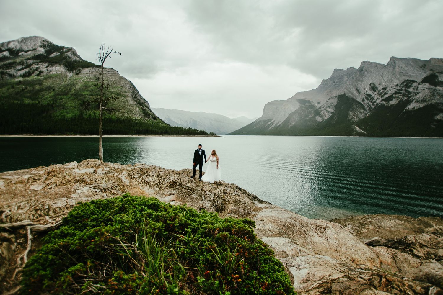 Two silhouettes stand on a rocky overlook beside a serene mountain lake surrounded by dramatic peaks in moody weather.