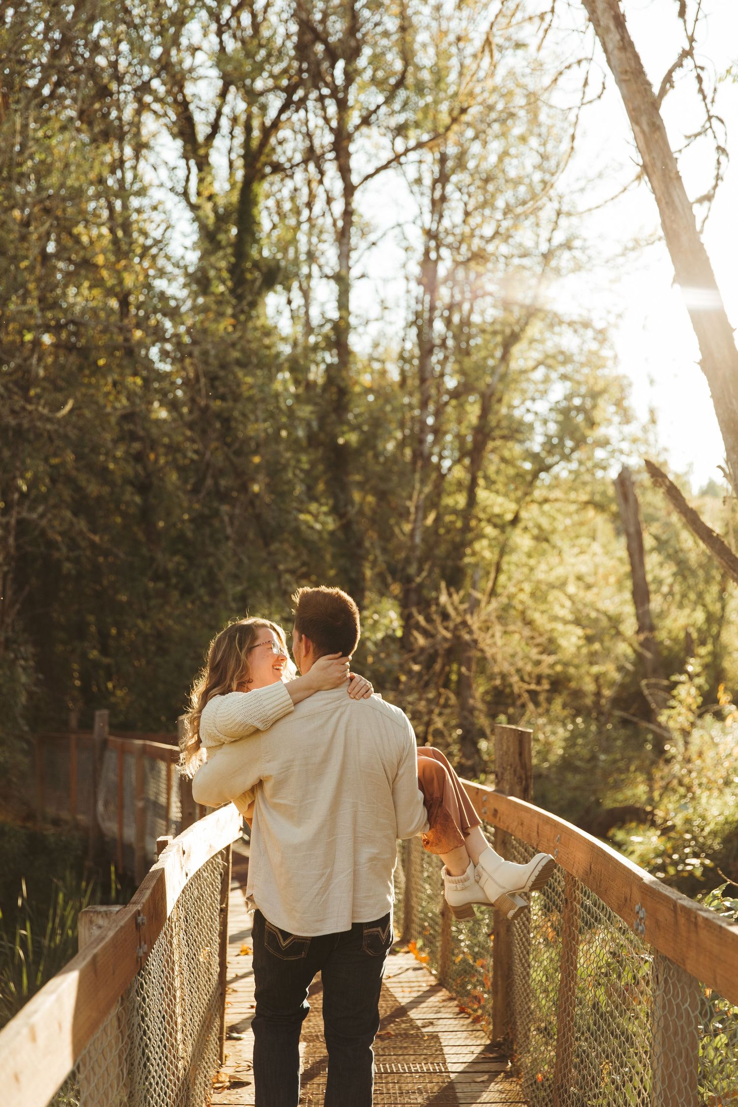 A couple in vintage attire dances romantically on a wooden bridge surrounded by sunlit trees at golden hour.