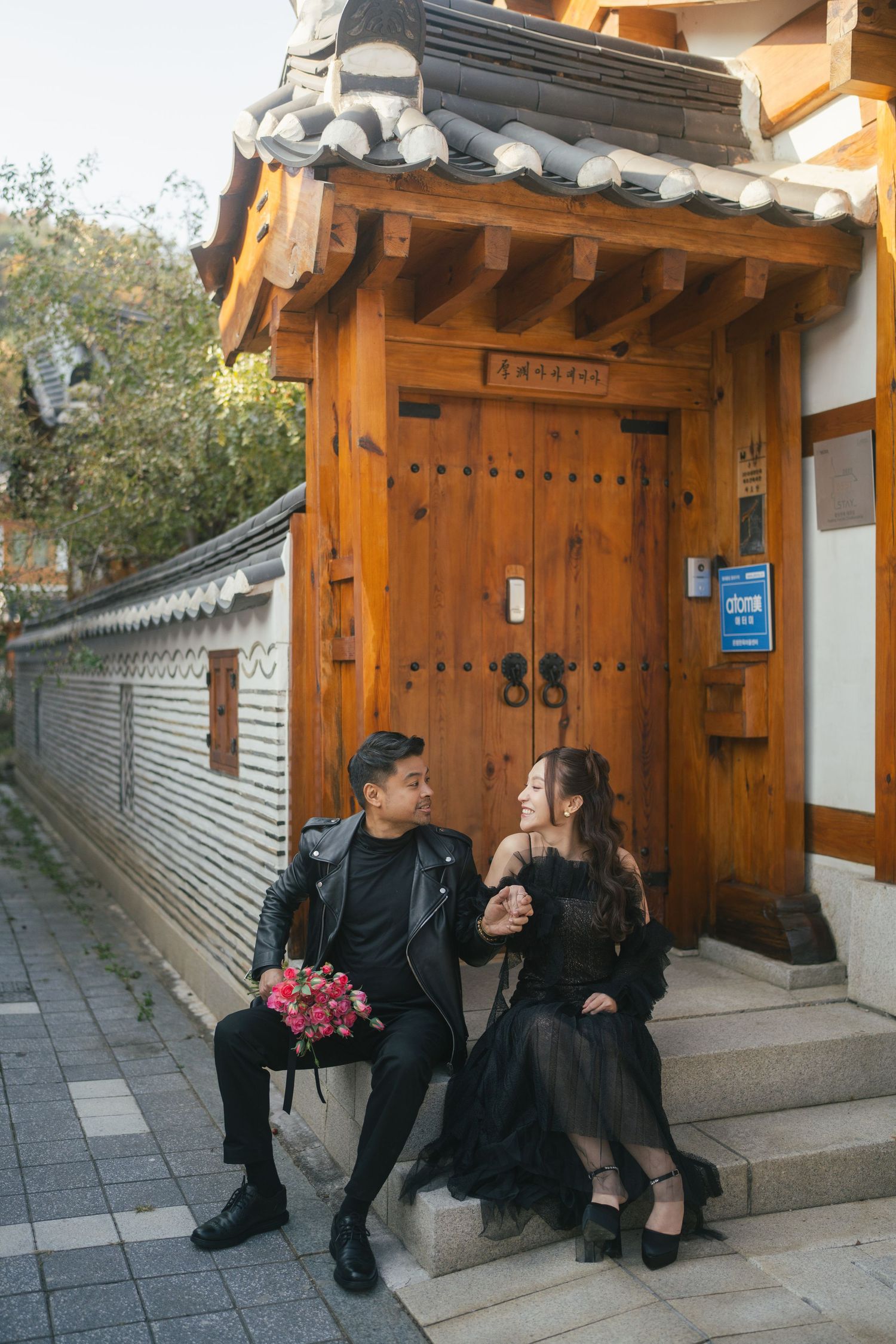 Couple sitting together along a traditional Korean hanok wall and pathway in matching black outfits.