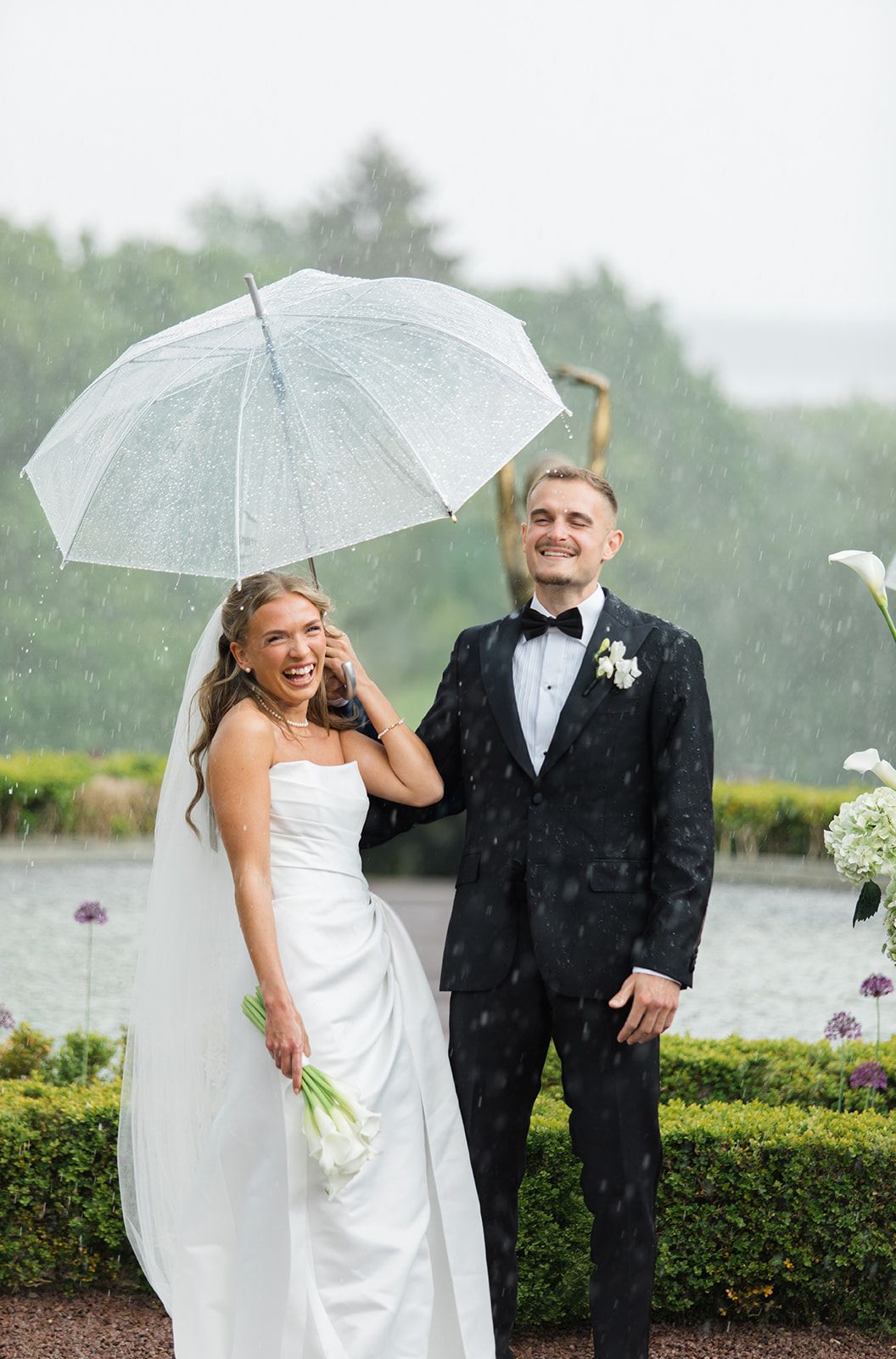 A couple shares a joyful moment in the rain under a white umbrella during their outdoor wedding celebration.