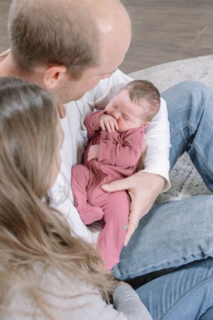Parents cuddling with newborn baby wearing pink outfit on gray couch.