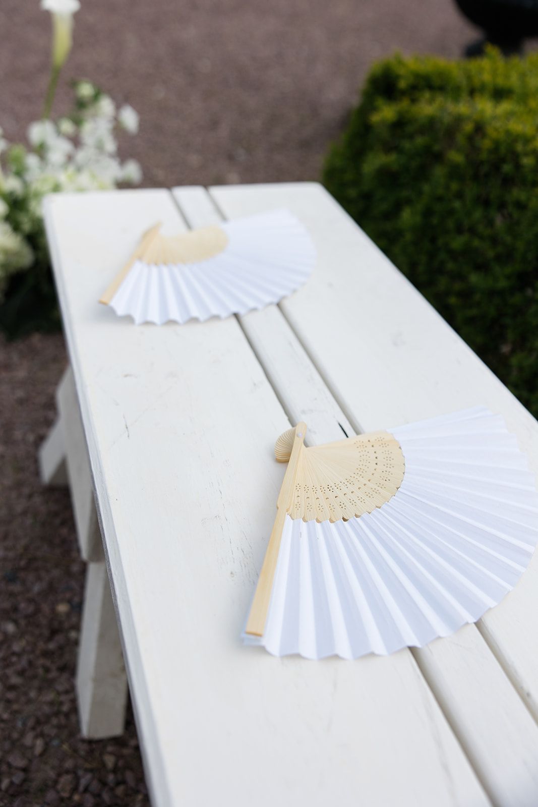 White paper hand fan resting on a white wooden bench in an outdoor garden setting.
