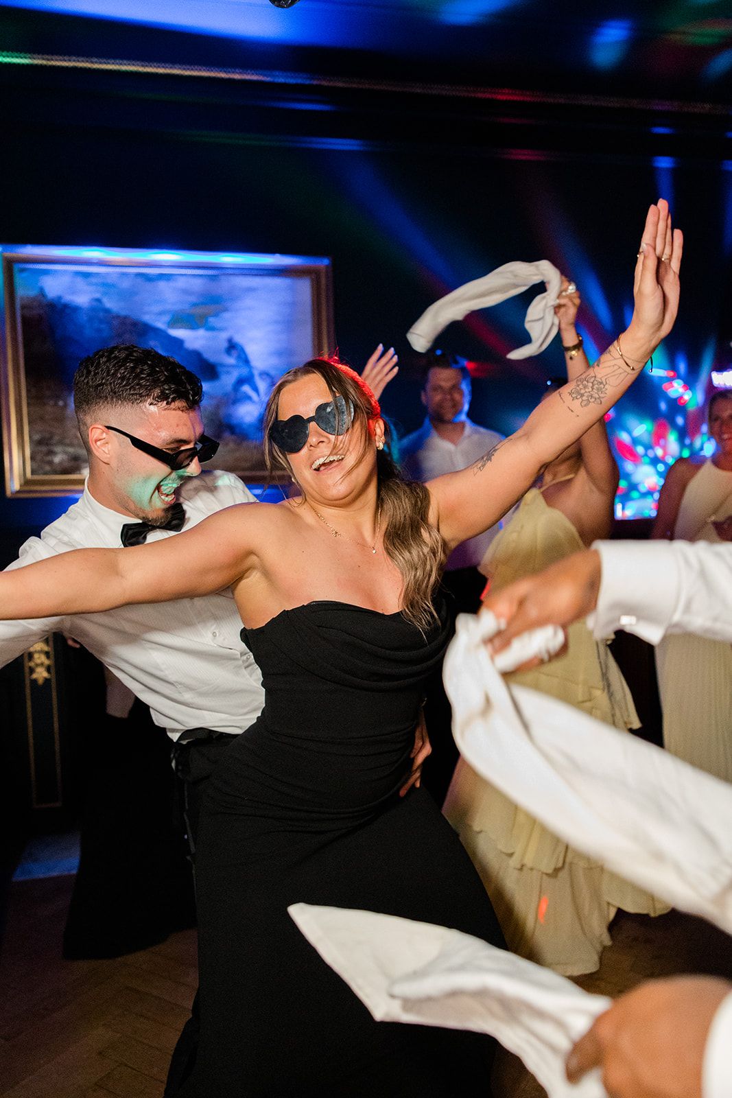 Wedding guests dance energetically at a reception with blue lighting and colorful effects in the background.