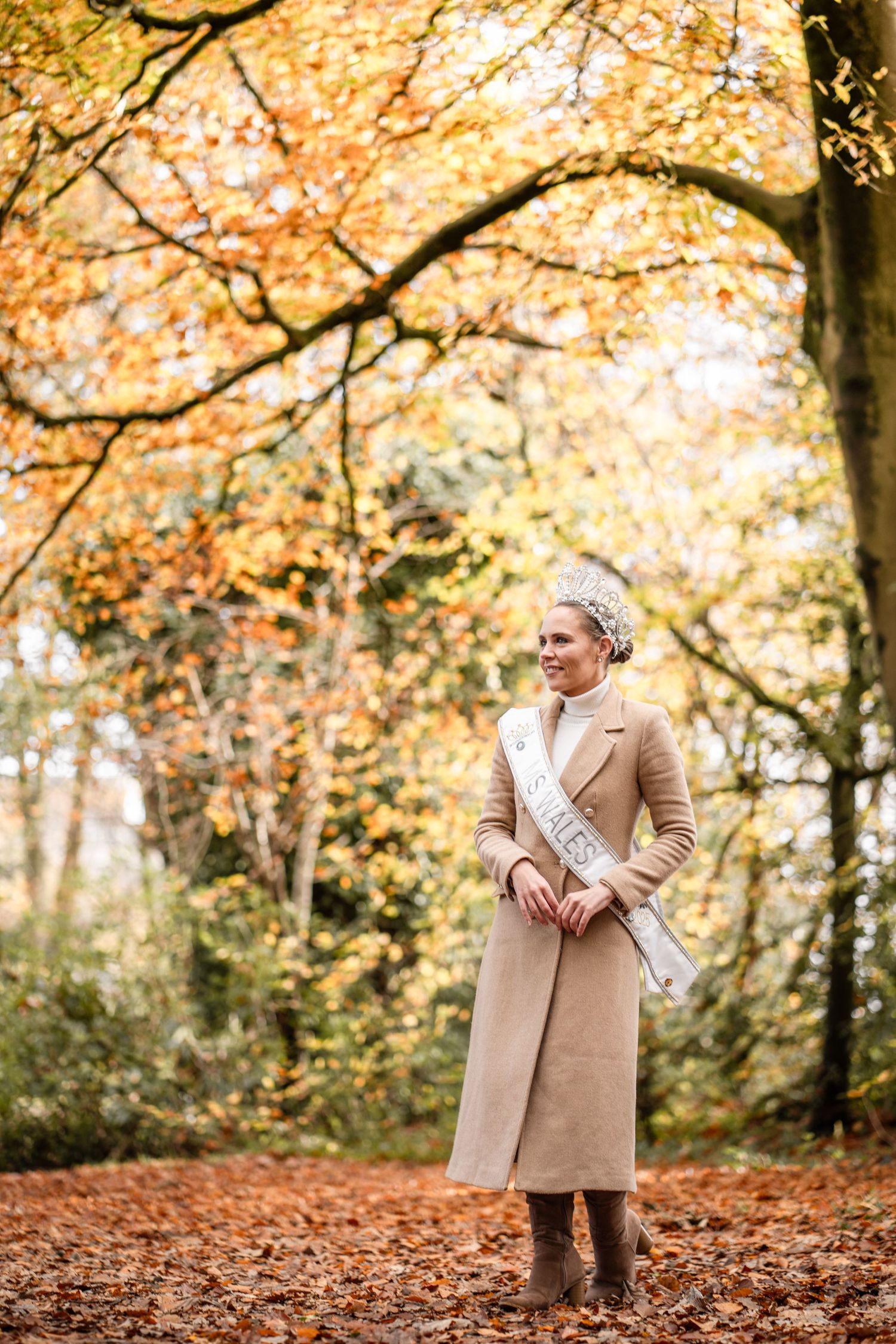 A person wearing a beige coat stands in a forest surrounded by autumn leaves and trees with golden foliage.