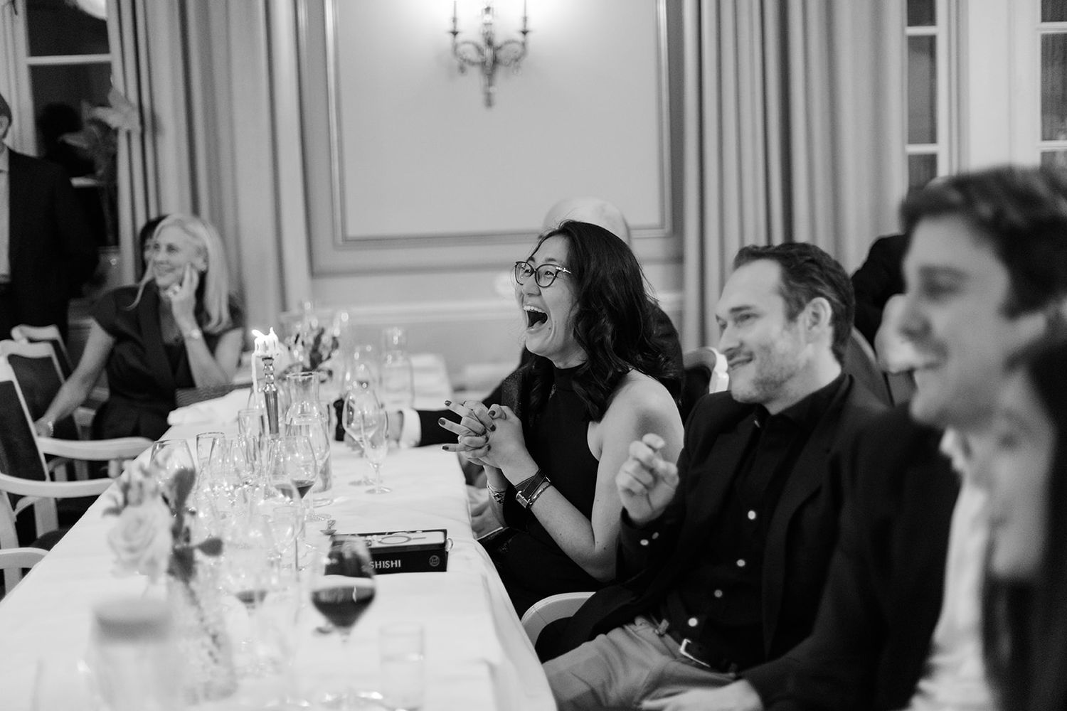 A group of people gathered around elegant dining tables at a formal event with white tablecloths and wine glasses.