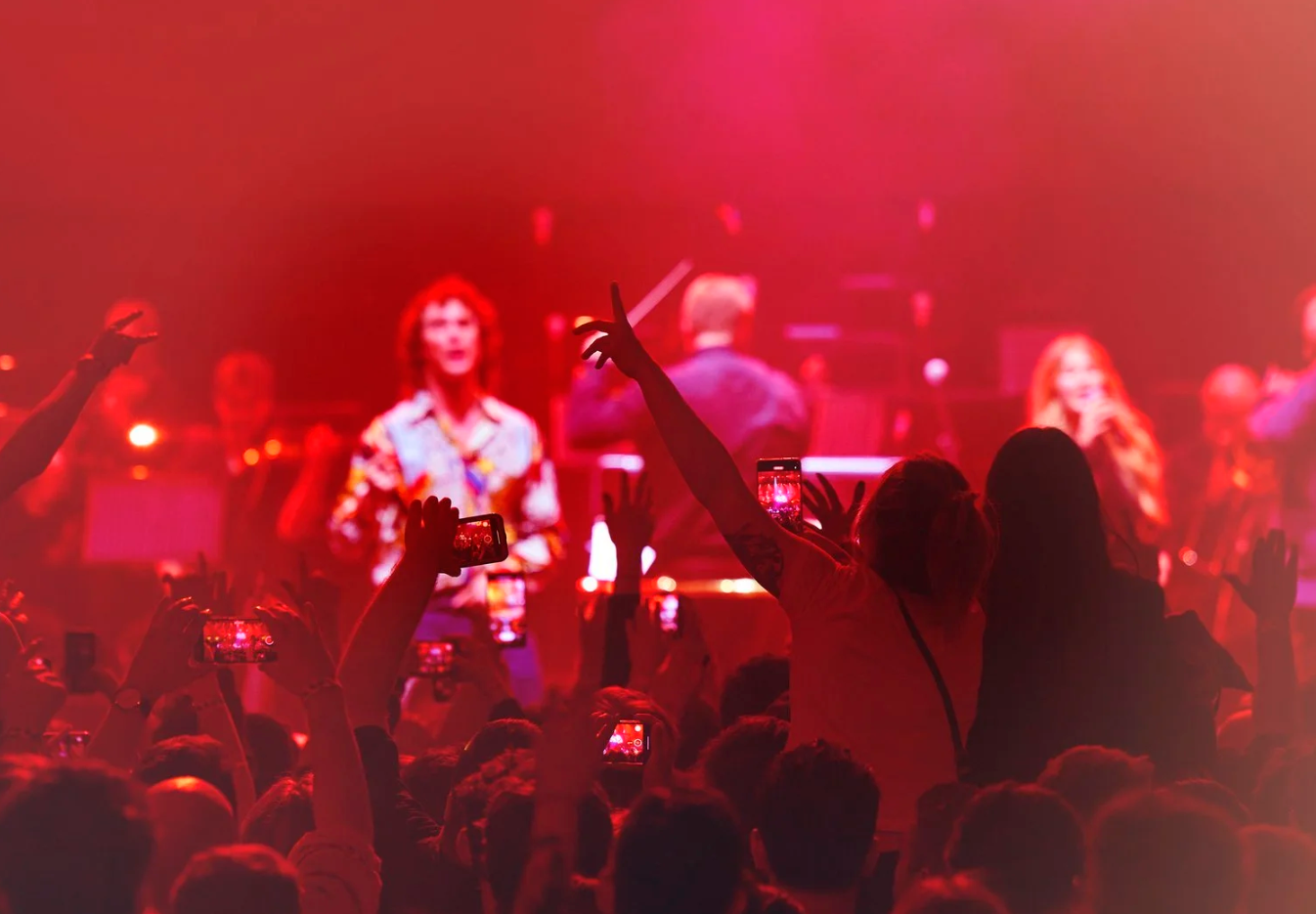Concert crowd silhouettes raise hands as performers play on stage bathed in dramatic red lighting.