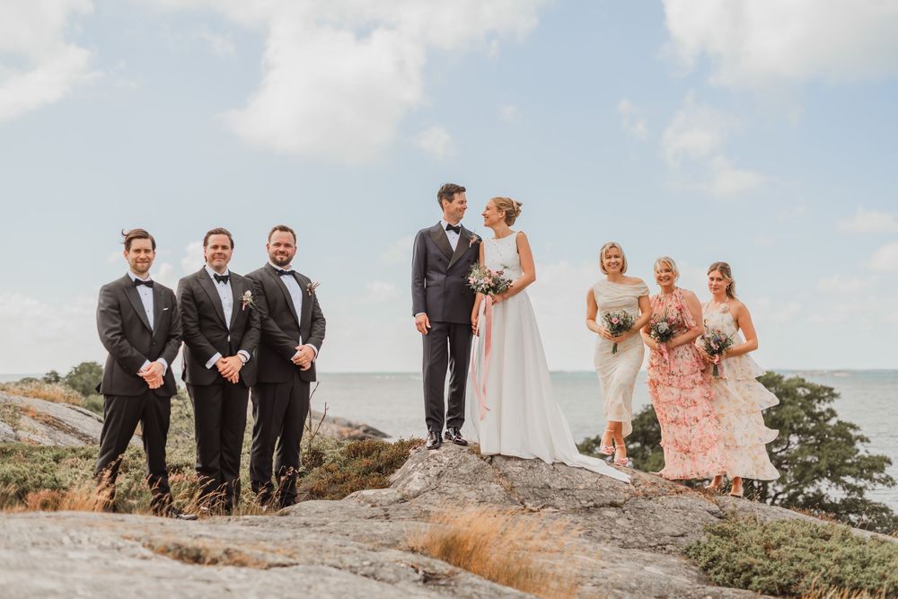 Wedding party poses on a rocky cliff overlooking a scenic coastal landscape during sunset.