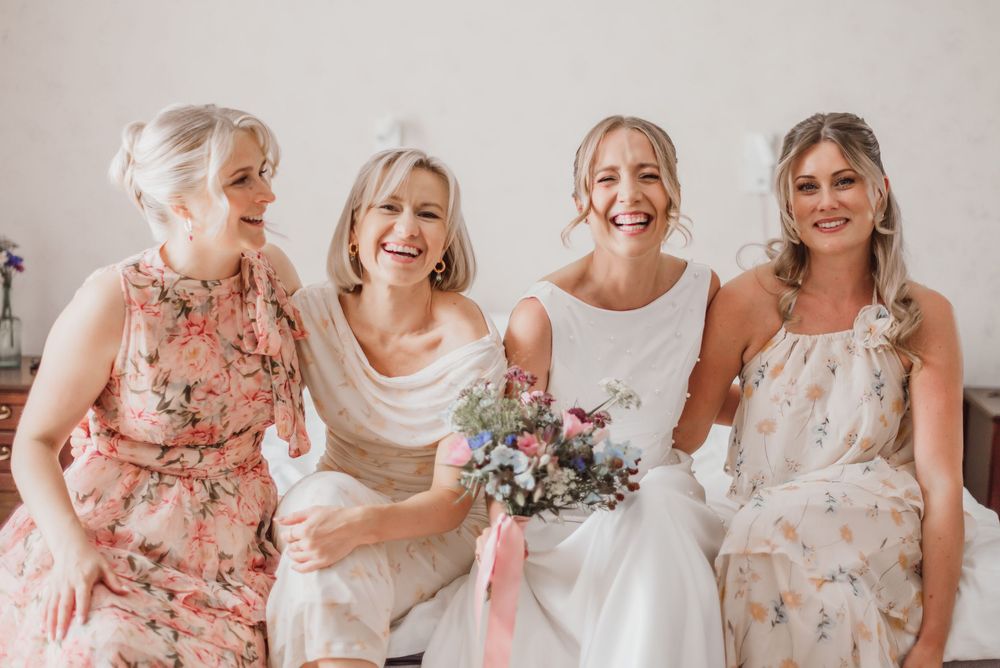 Bridesmaids in floral pink dresses share joyful laughter with bride in white gown during wedding preparations.