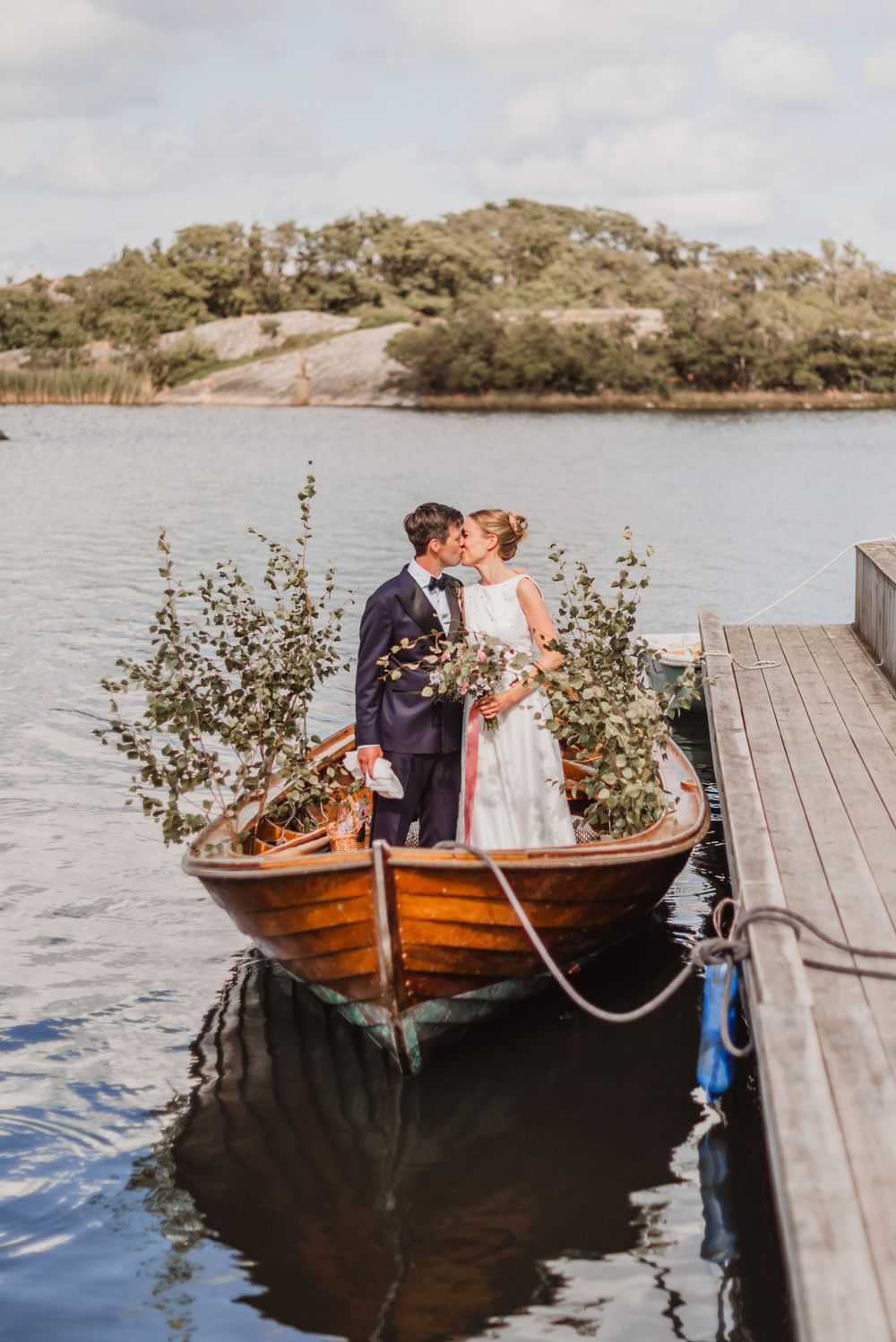 A romantic couple shares tender moments in a wooden rowboat by a dock on a serene lake.