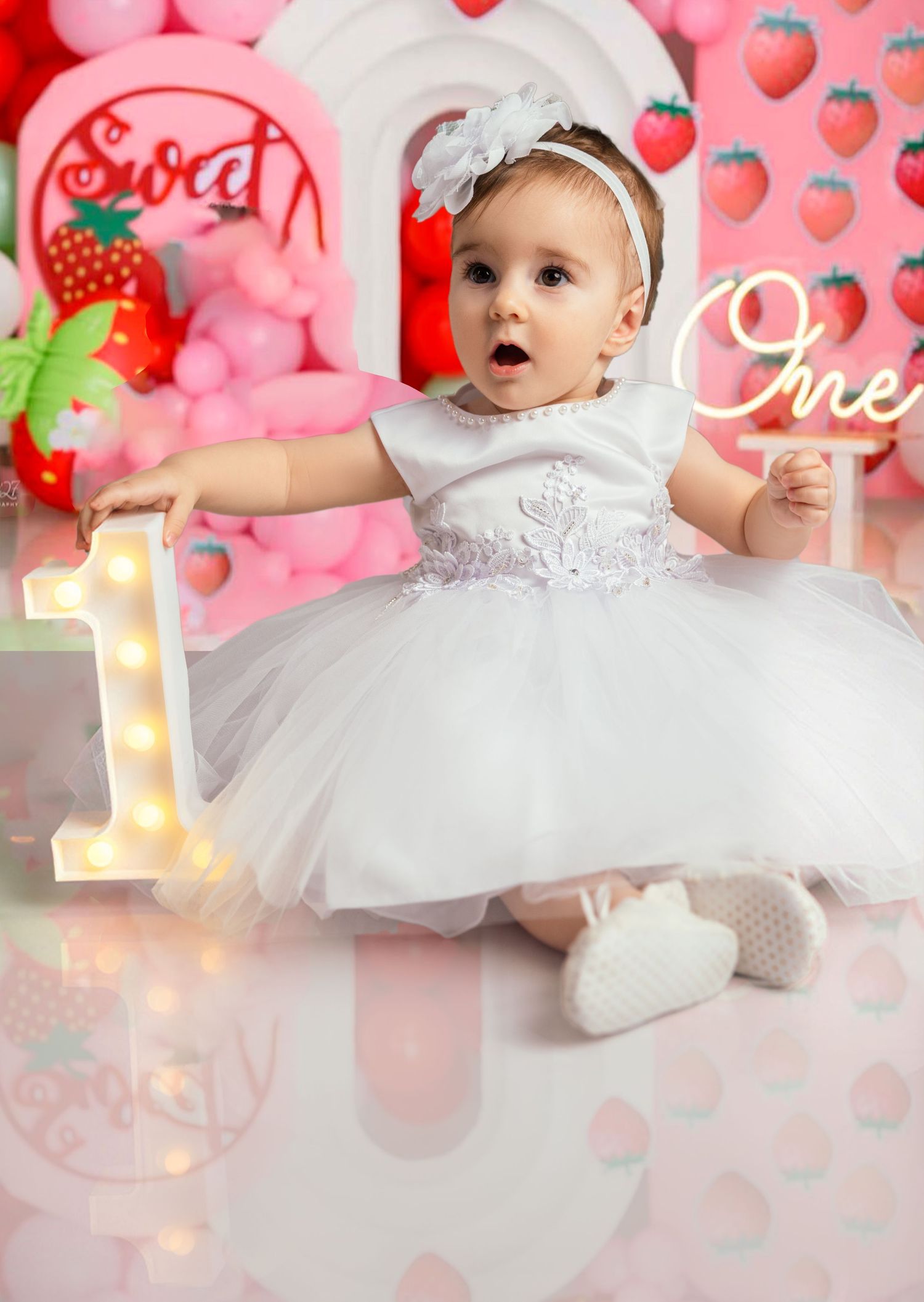 Baby girl in white tutu dress celebrating first birthday with strawberry themed decor.