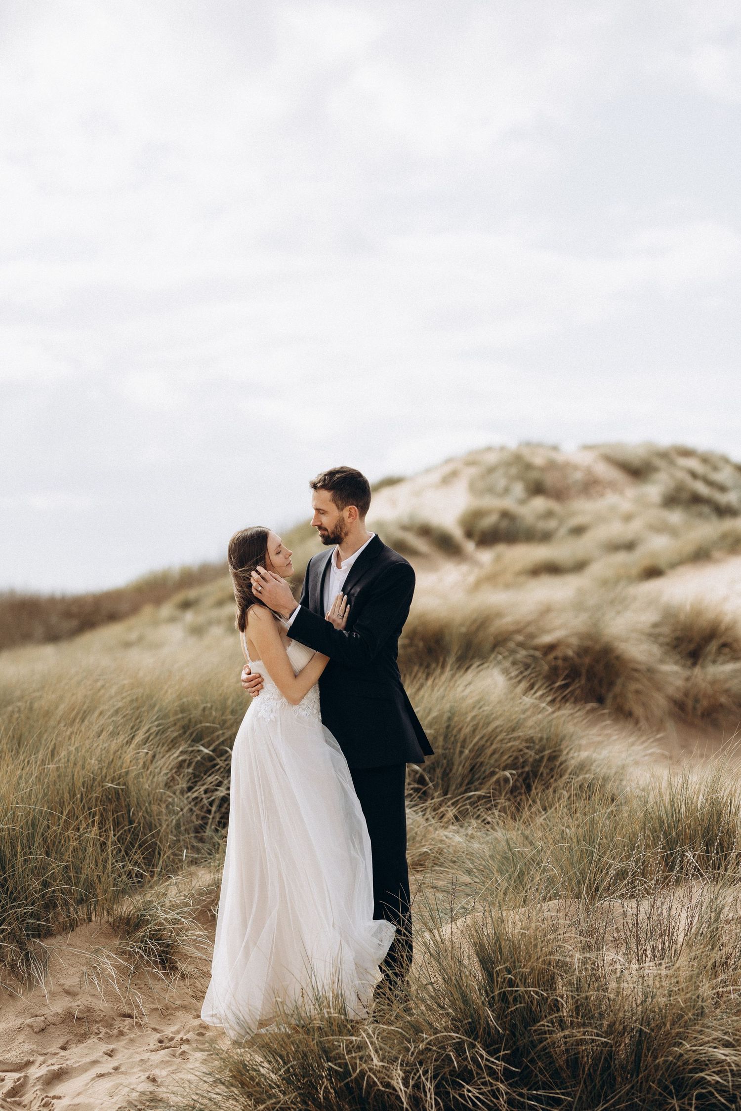 A couple embraces on a windswept coastal dune surrounded by tall grass and cloudy skies.