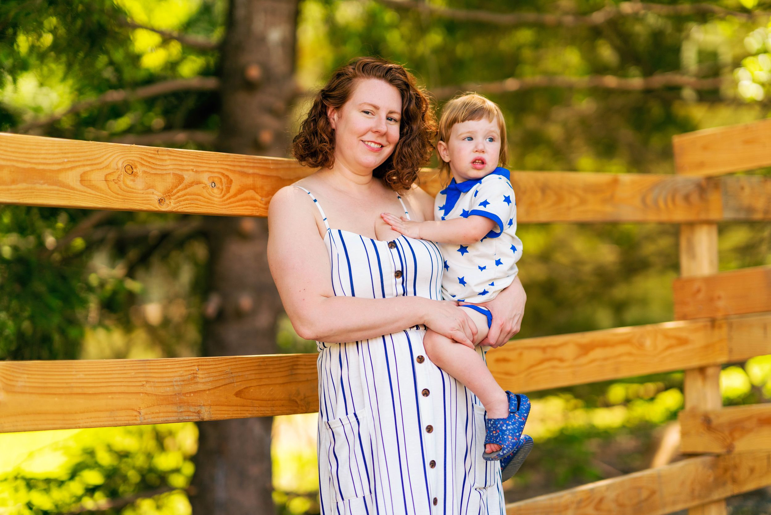 A woman in a striped dress holds a baby in blue while standing on a rustic wooden bridge in a sunny forest setting.