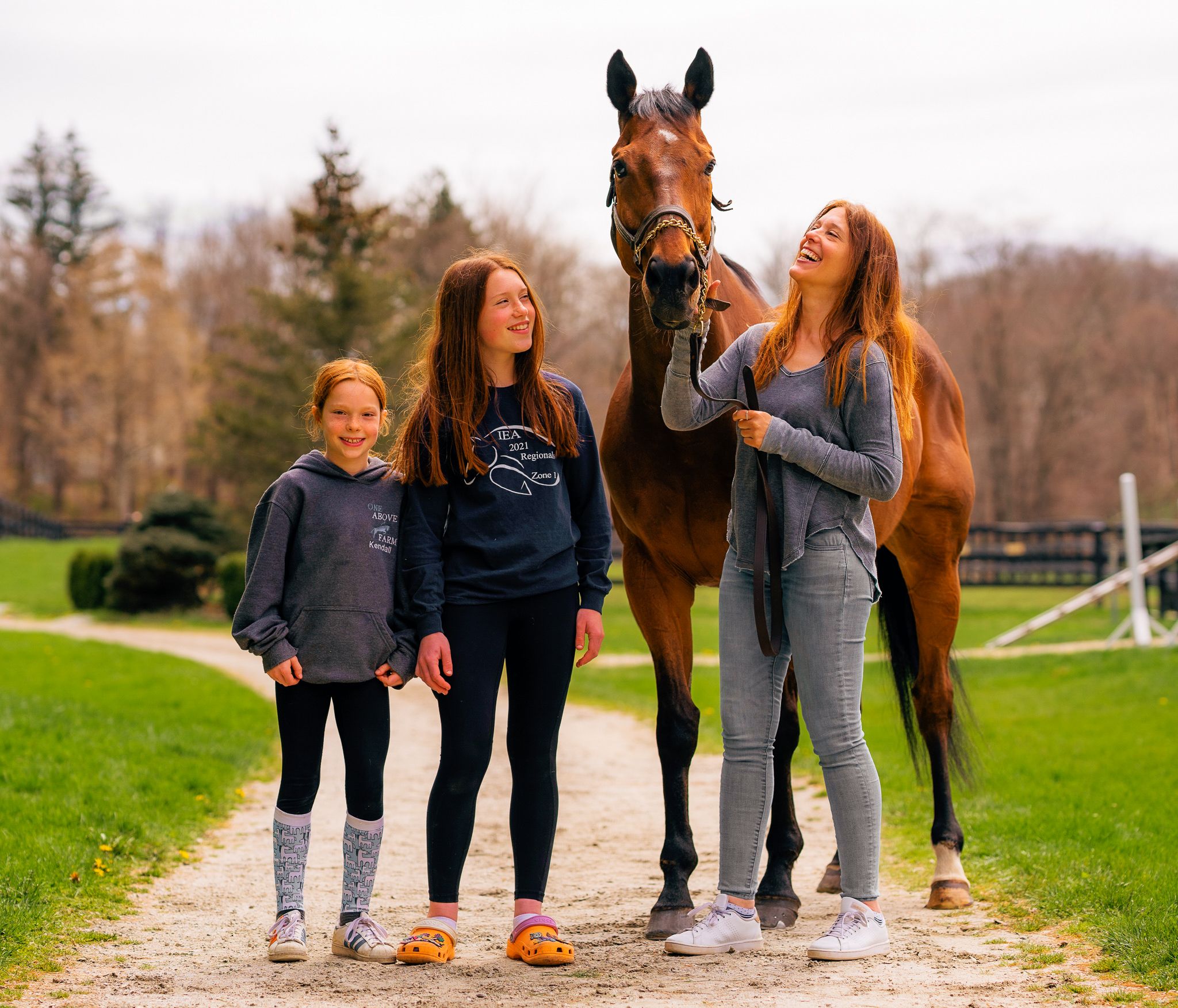 Group posing with beautiful bay horse on dirt path with fenced paddock in background.
