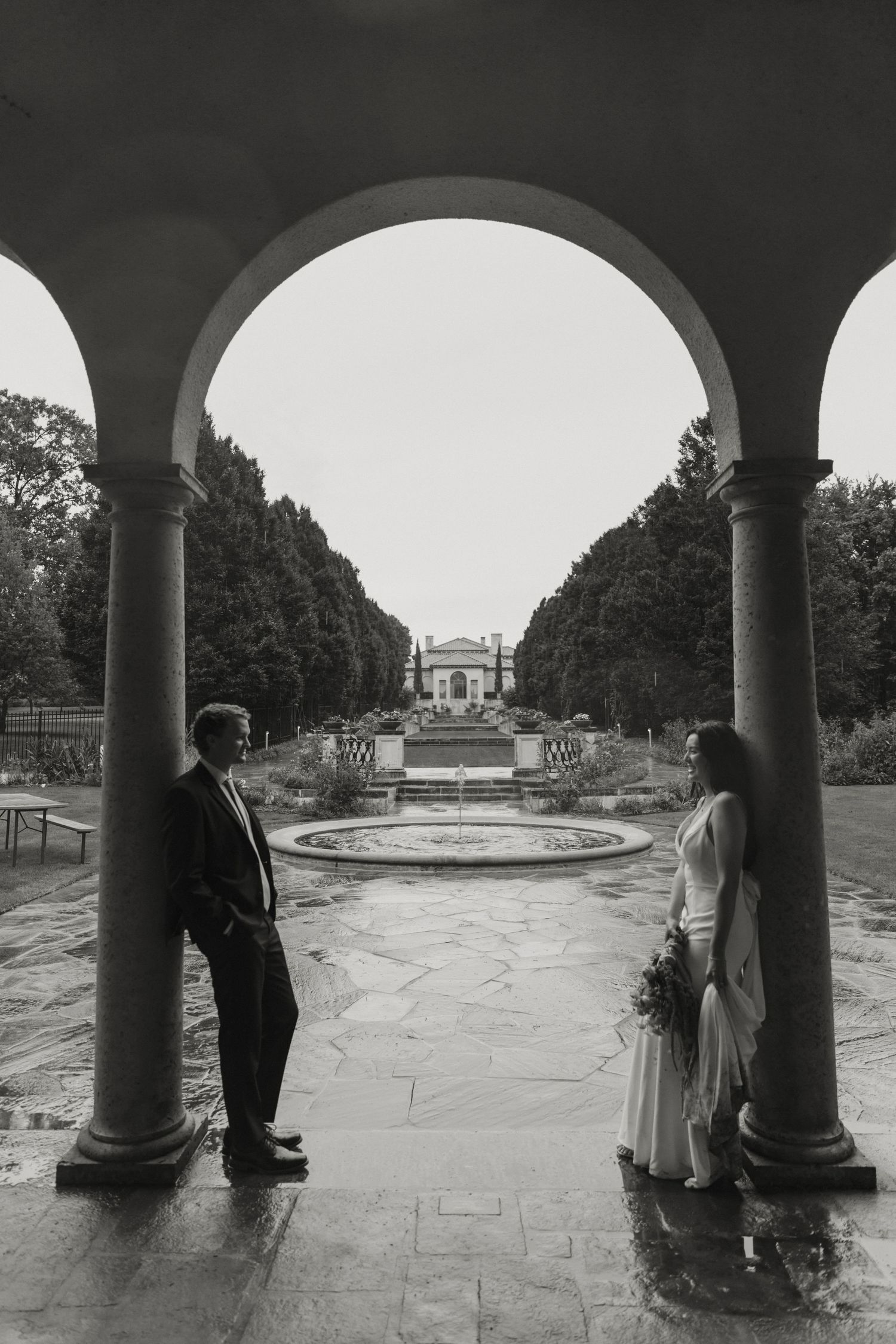 Two people stand beneath arches overlooking a formal garden with fountains and a classical building in the distance.