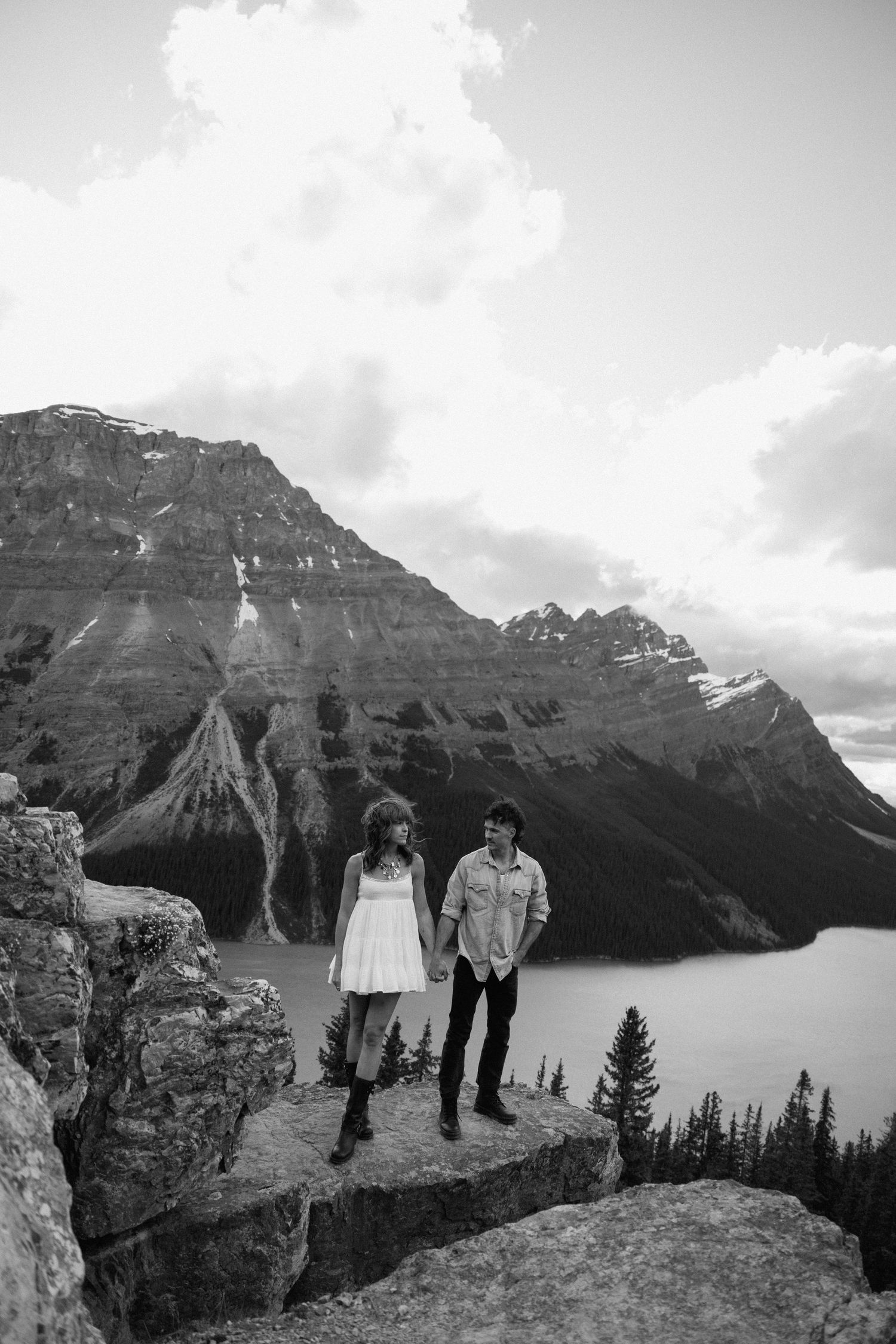 A couple poses together in a scenic mountain meadow filled with vibrant wildflowers and dramatic rocky peaks.