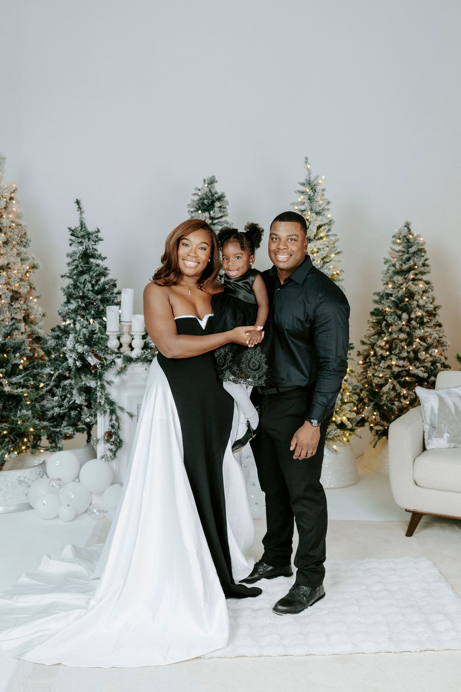A couple in formal attire poses playfully in front of decorated Christmas trees in a white studio setting.