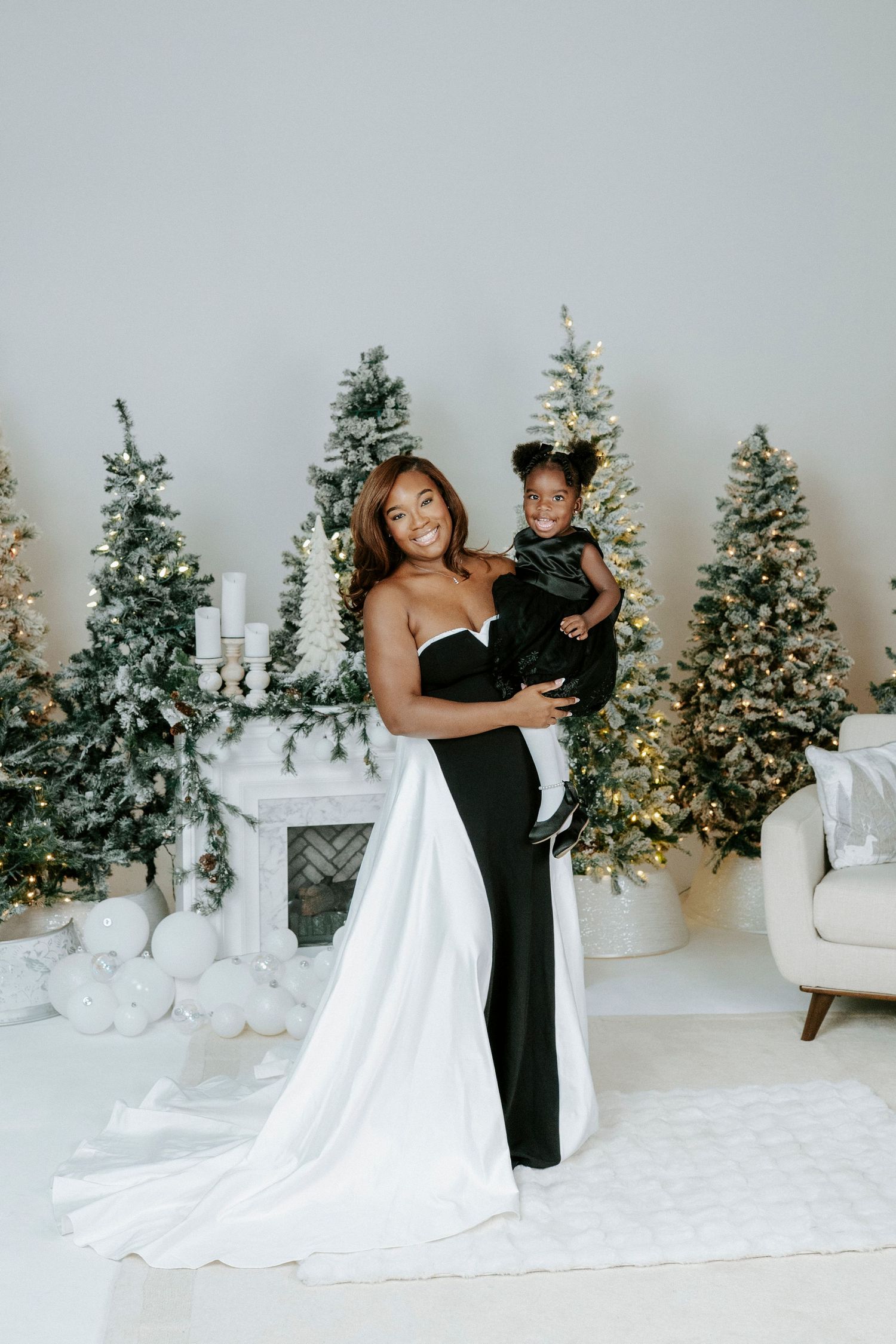A couple in formal attire embraces in front of illuminated Christmas trees in an all-white studio setting.