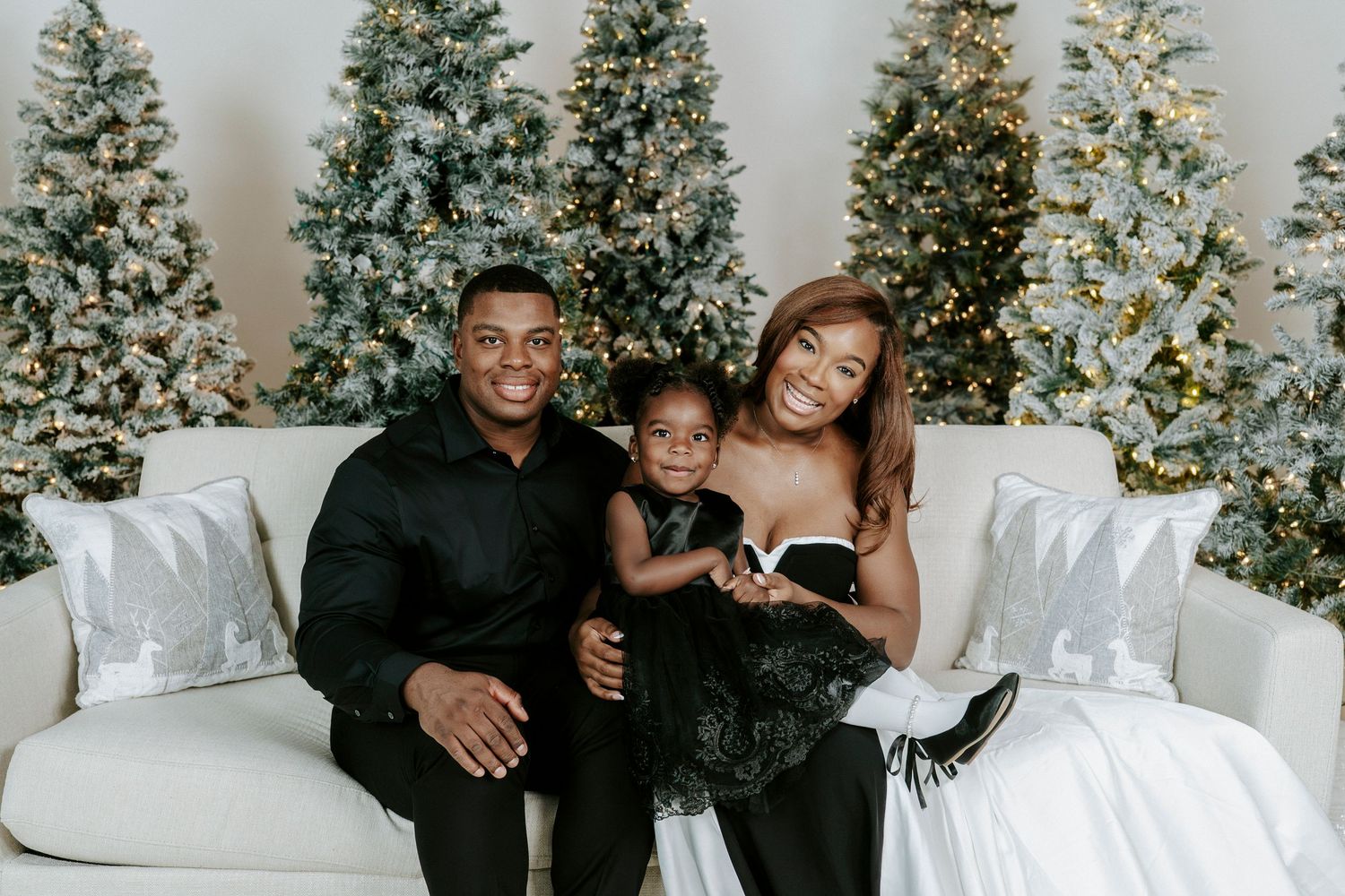 Family poses together for holiday photos in front of decorated Christmas trees in a bright white studio setting.