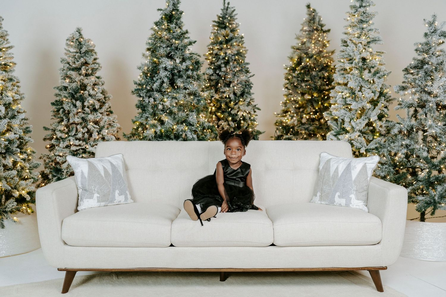 A white modern sofa sits in front of decorated Christmas trees with twinkling lights in a bright interior setting.