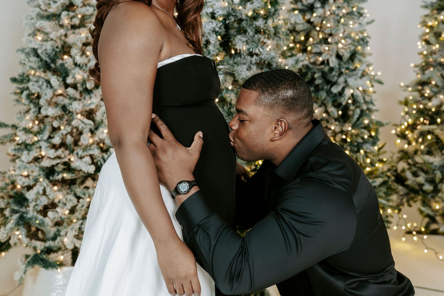 A romantic pregnancy announcement photo in front of decorated Christmas trees with twinkling lights.