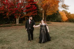 A couple walks together on a grassy hill surrounded by autumn trees.