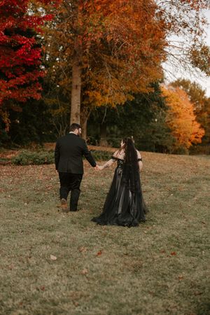 A couple strolls hand in hand across a grassy slope with fall foliage in the background.