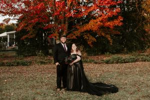 A couple poses for photos on a lawn with vibrant fall foliage behind them.