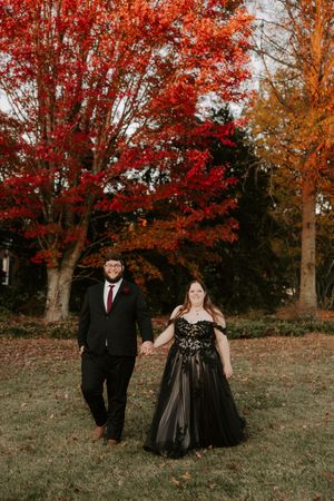 A couple holds hands while walking through a scenic fall landscape with bright red foliage.