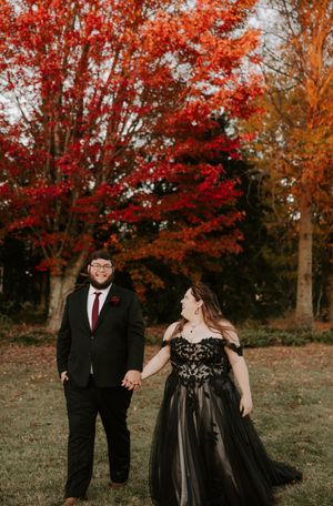 A couple in formal black attire enjoys a playful moment amid stunning fall colors.