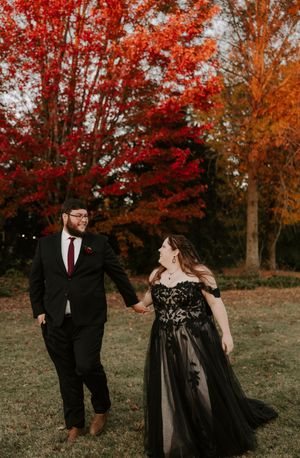 Couple in formal black attire dances and laughs together under vibrant autumn trees.