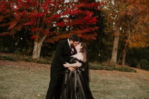 A couple in formal attire share a romantic moment against vibrant red autumn foliage in a park setting.