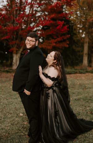A joyful couple in formal black attire dance together during their autumn outdoor photoshoot.