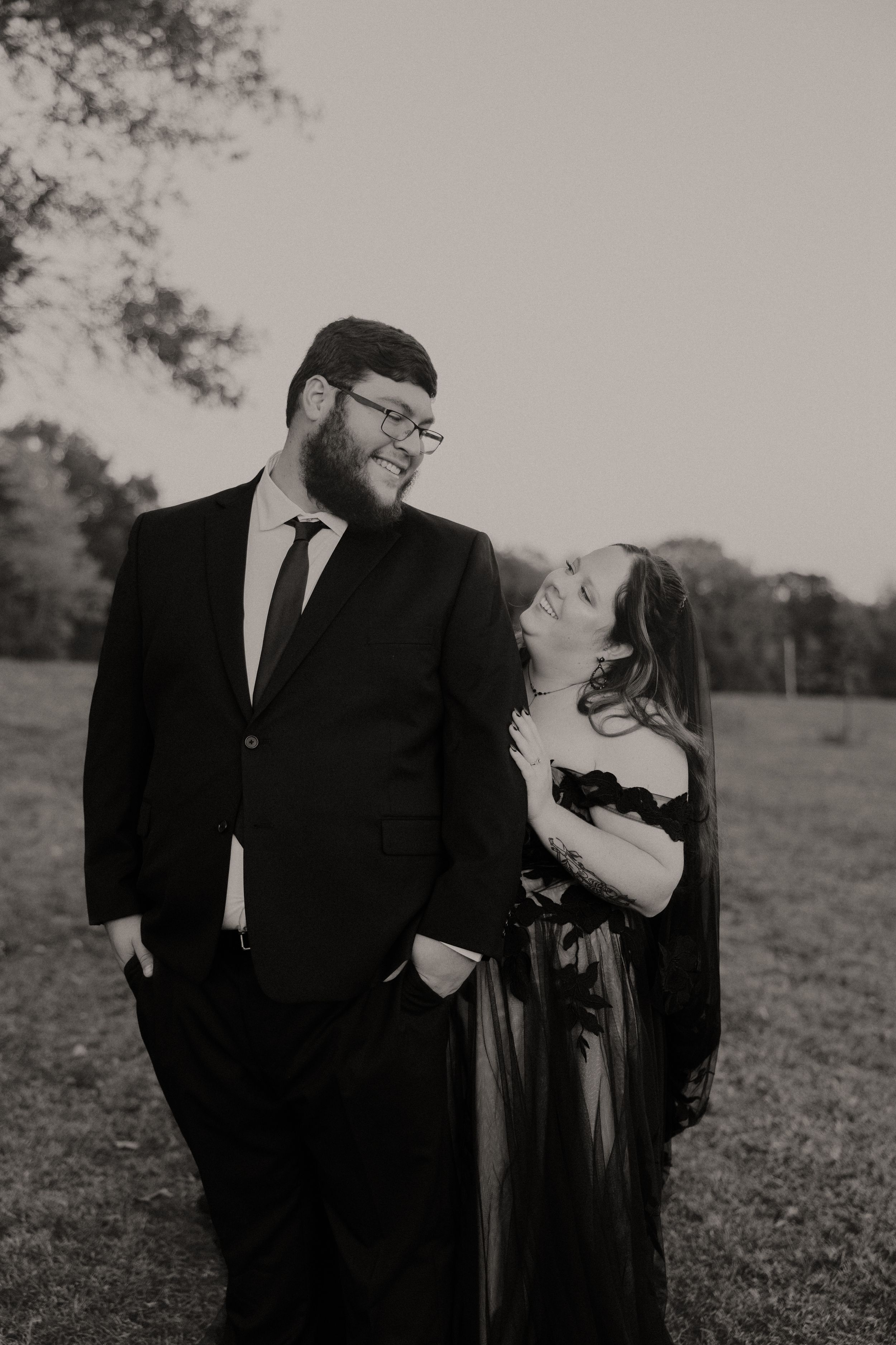 A black and white portrait of a couple in formal wear sharing a playful moment outdoors.
