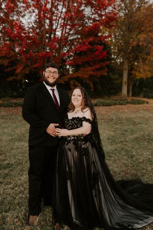A couple in formal black attire poses together on a hillside with stunning fall colors.