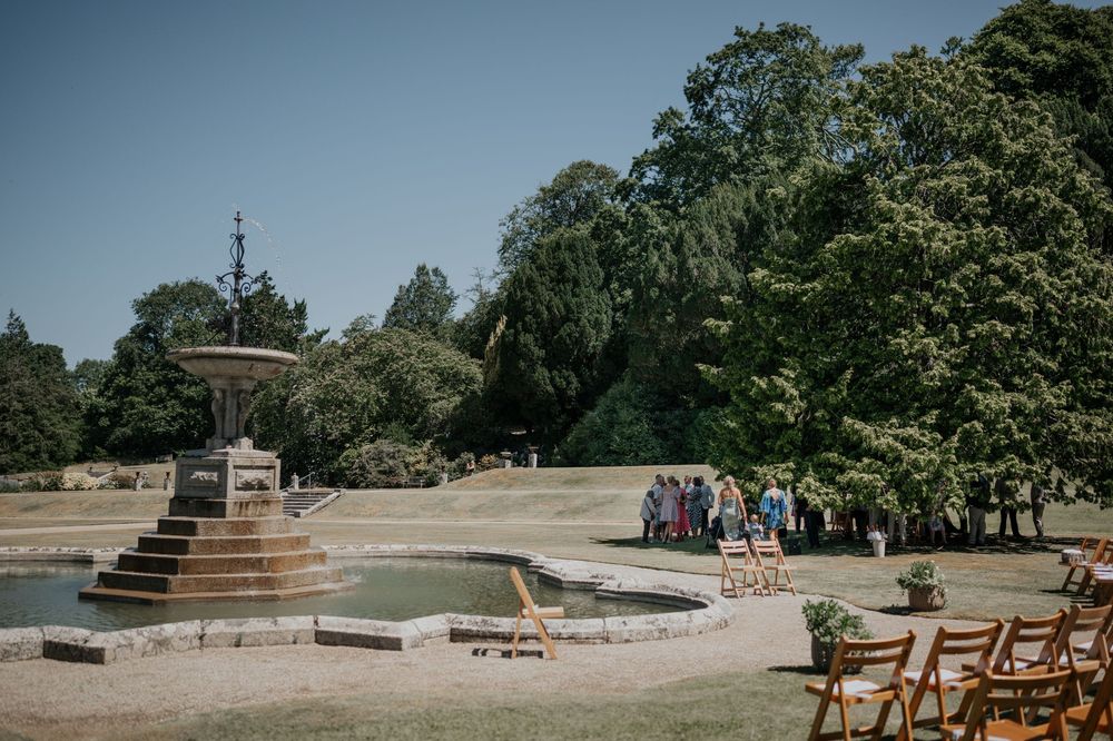 Guests gather near an ornate stone fountain and pond in the formal gardens of a grand country house wedding venue.