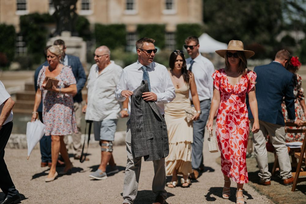Elegantly dressed wedding guests walking across a grand estate courtyard on a sunny day.