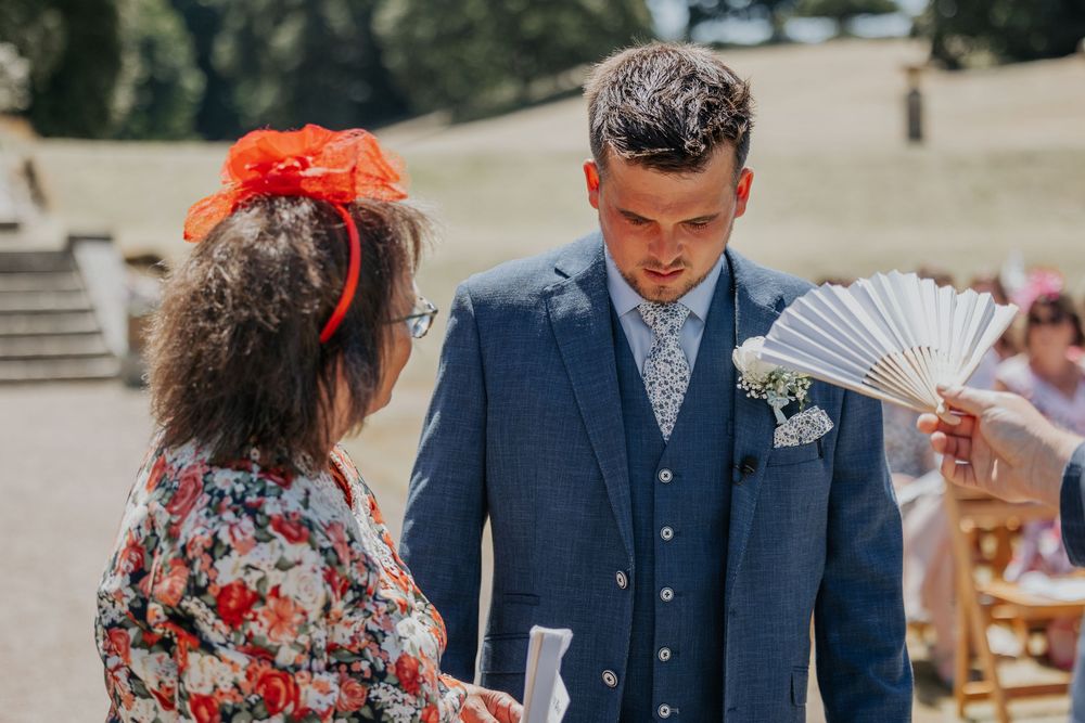 Groom in blue suit receiving a hand fan from a guest at a sunny outdoor wedding ceremony.