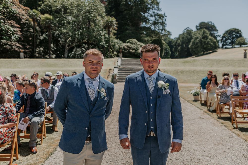 Two groomsmen in matching blue suits walking down the aisle at a sunny outdoor wedding ceremony.