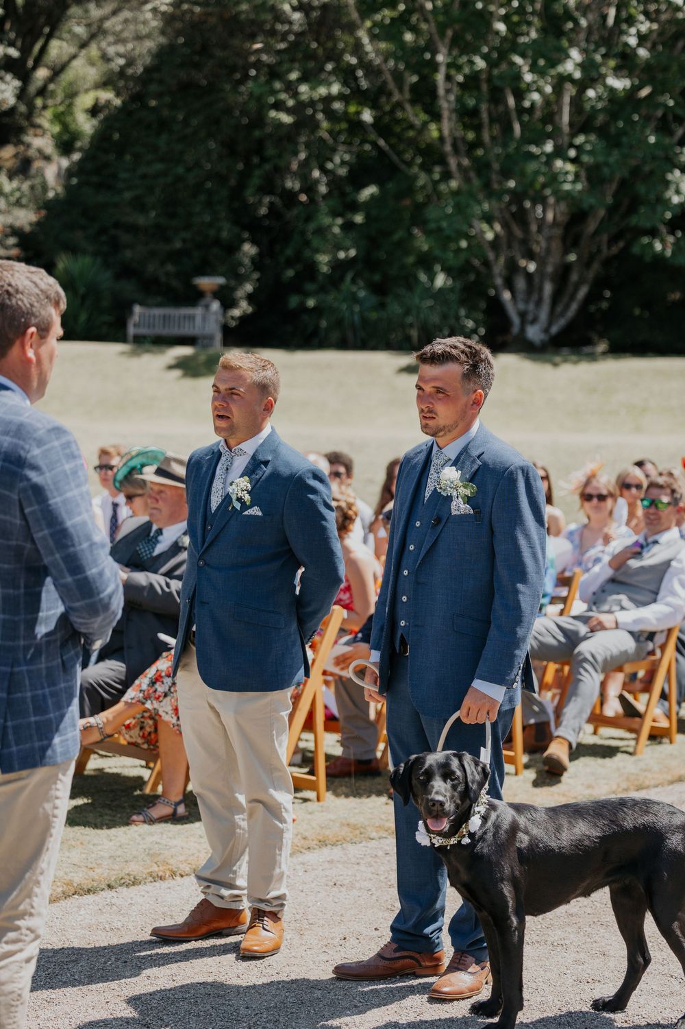 Two groomsmen in blue suits standing at the altar with a black Labrador dog at an outdoor wedding.