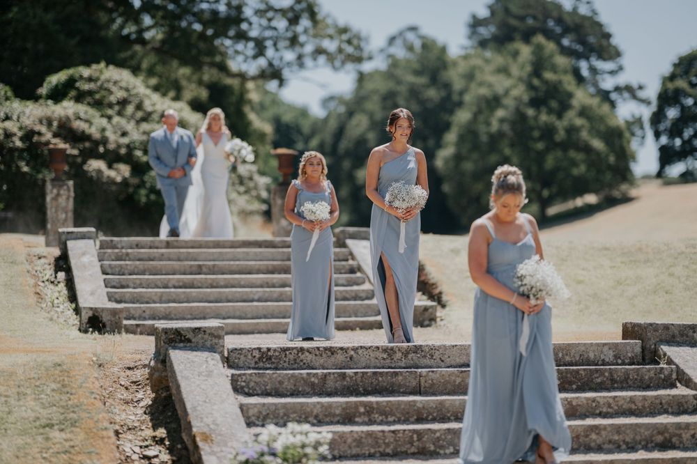 Bridesmaids in blue gowns and bride with father descending grand stone steps at a wedding.