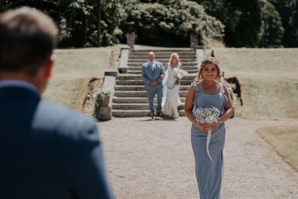 Bridesmaid in blue dress descending stone steps carrying a bouquet at an outdoor wedding.