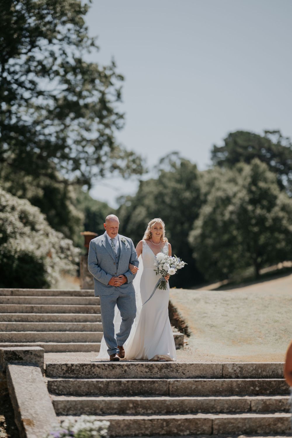 Bride in elegant white gown walking down stone steps with her father on a sunny wedding day.