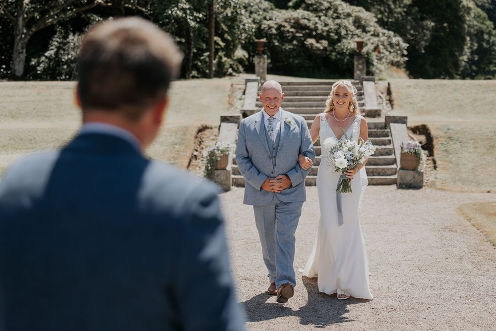 Bride walks down outdoor aisle with father toward groom at elegant garden wedding ceremony.