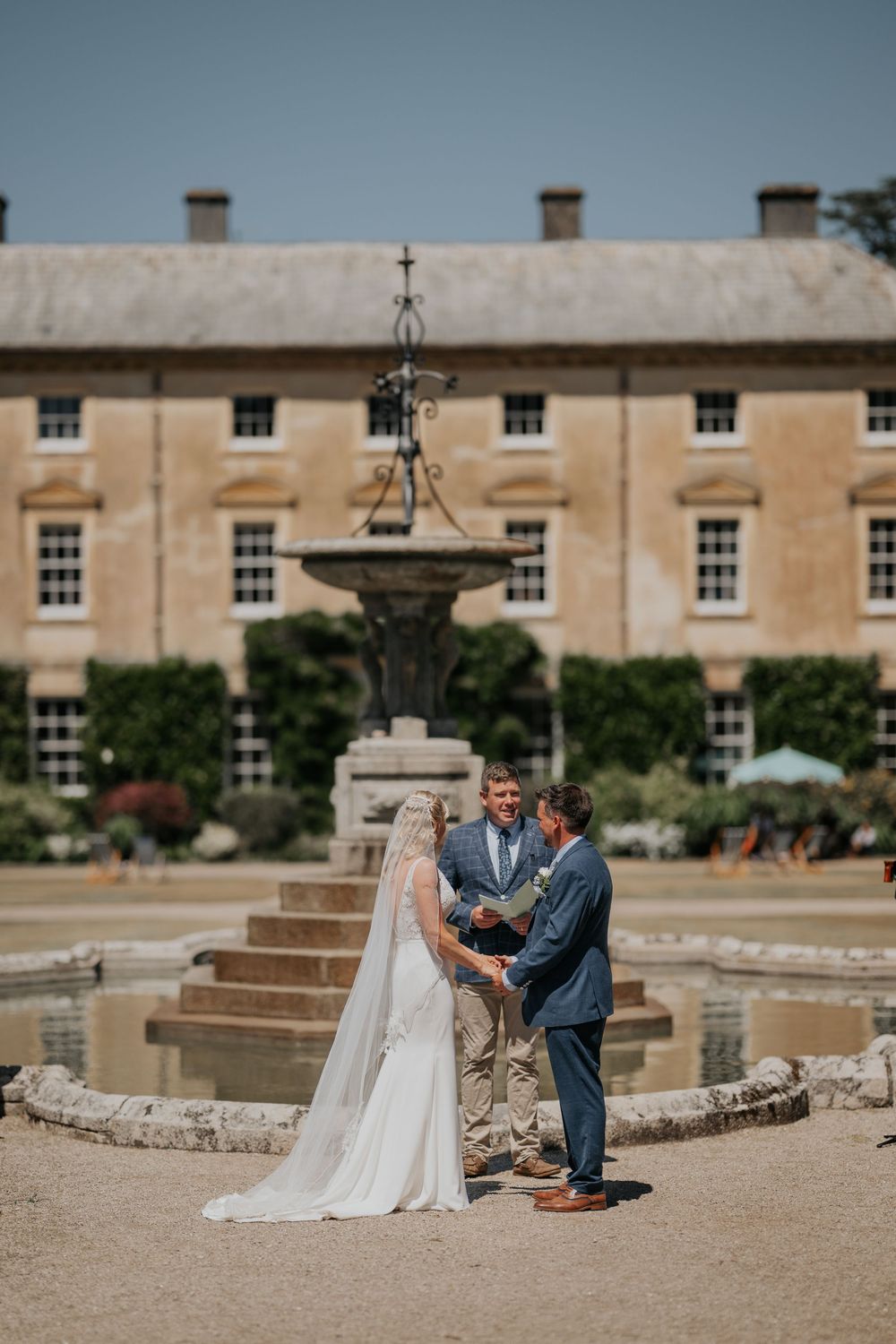 Bride and groom exchange vows before officiant at outdoor wedding ceremony by ornate fountain.