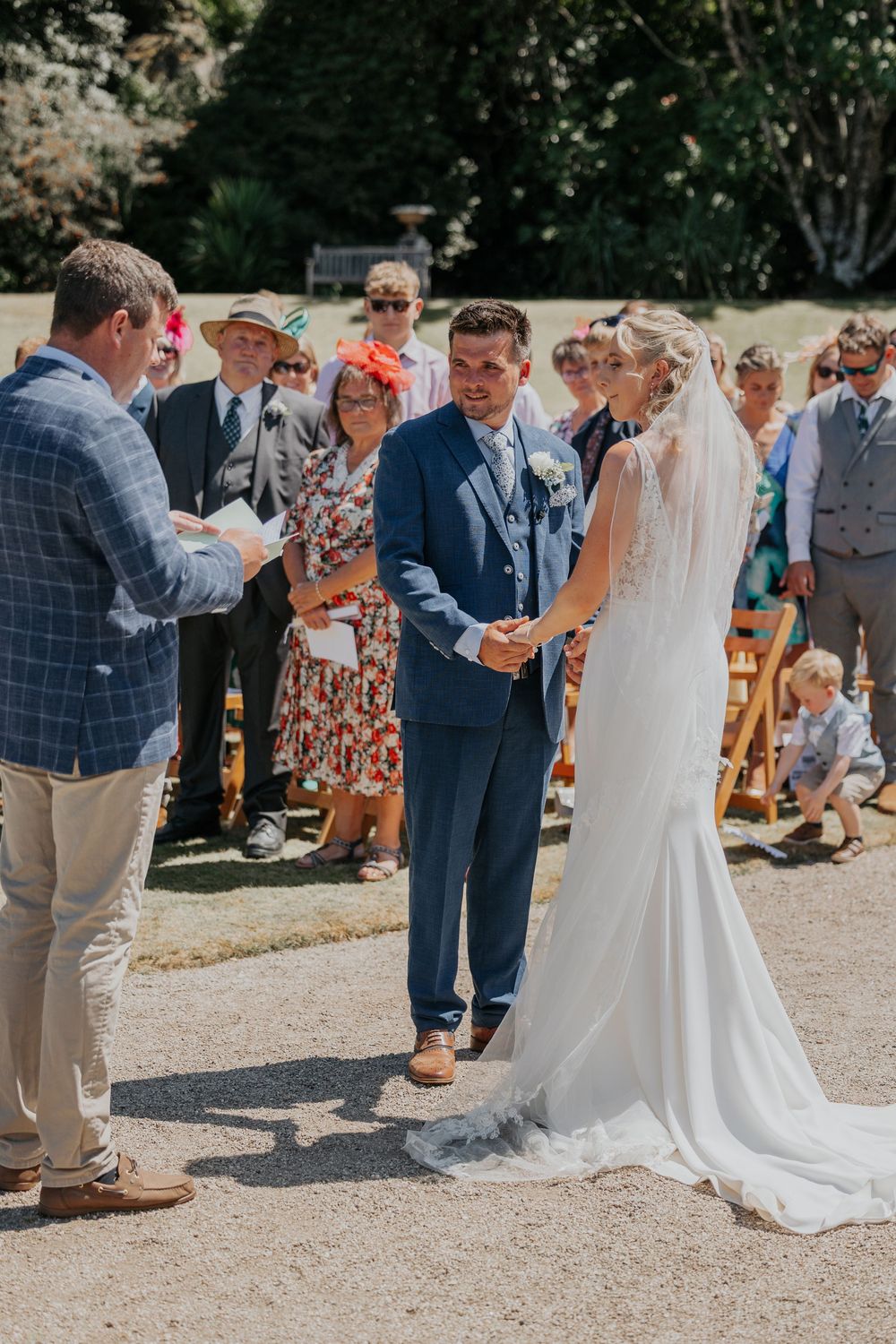Groom smiles at bride during outdoor wedding ceremony with seated guests in background.
