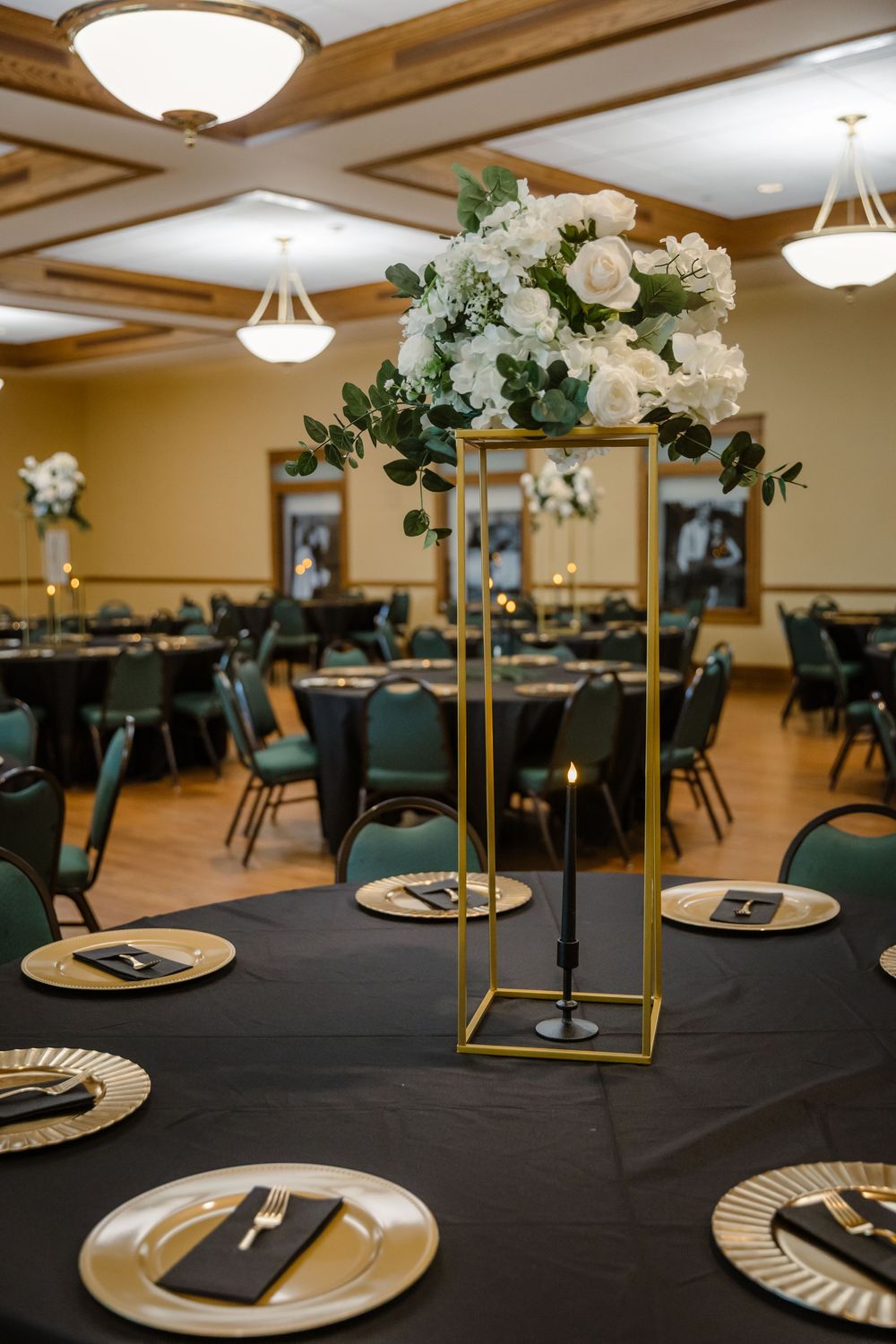 Elegant banquet hall setup with black tablecloths, gold stands, and white floral centerpieces in a warm-lit venue.